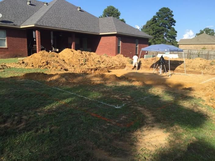 Construction workers dig a backyard pool foundation in front of a brick house on a sunny day.