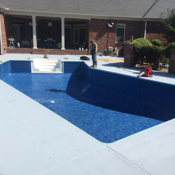 A worker stands by a freshly lined, deep blue swimming pool in a backyard with a brick house and light gray patio.