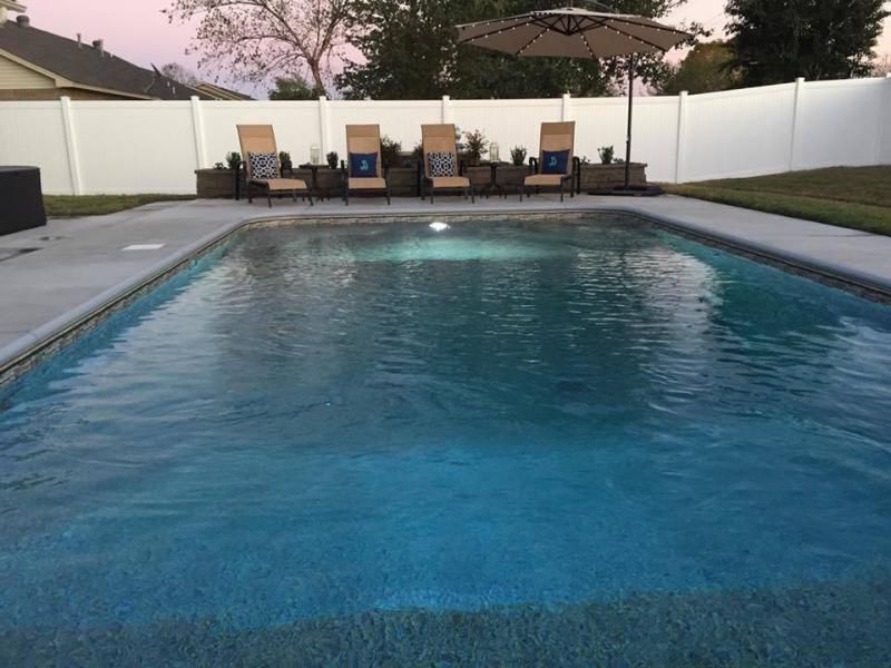 A backyard swimming pool with a stone deck, four lounge chairs under an umbrella, and a white privacy fence at dusk.