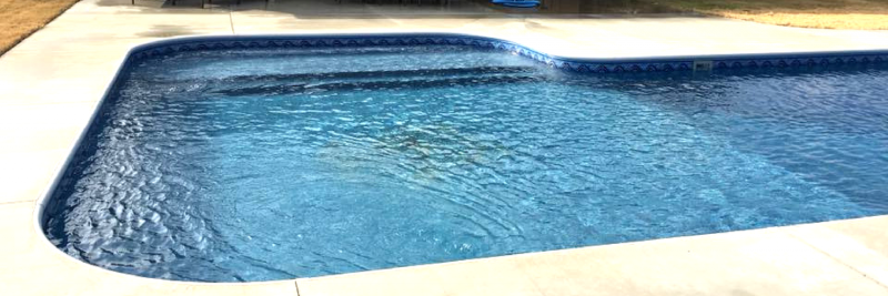 A blue rectangular swimming pool with rounded corners and a concrete deck under bright sunlight.