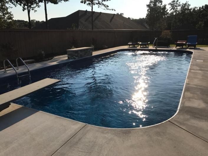 A rectangular swimming pool with deep blue water, a diving board, and a stone water feature on a concrete patio.