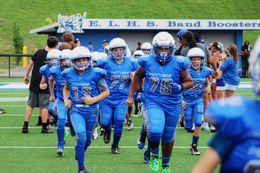 Blue-clad football team runs onto a field beneath a school banner, led by players in helmets and jerseys.