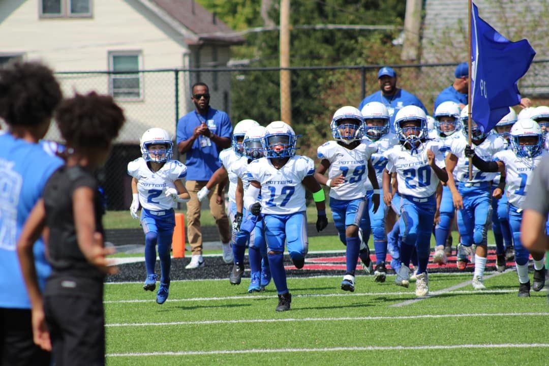 Youth football player in blue runs between cheerleaders on a green field.
