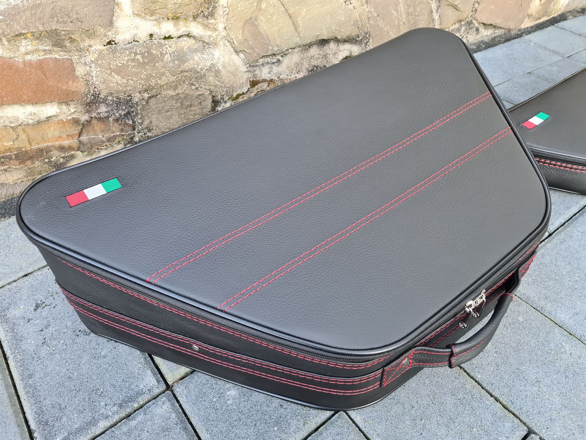 Een zwarte aktetas met rode stiksels en een handvat staat op een stoep van bakstenen.