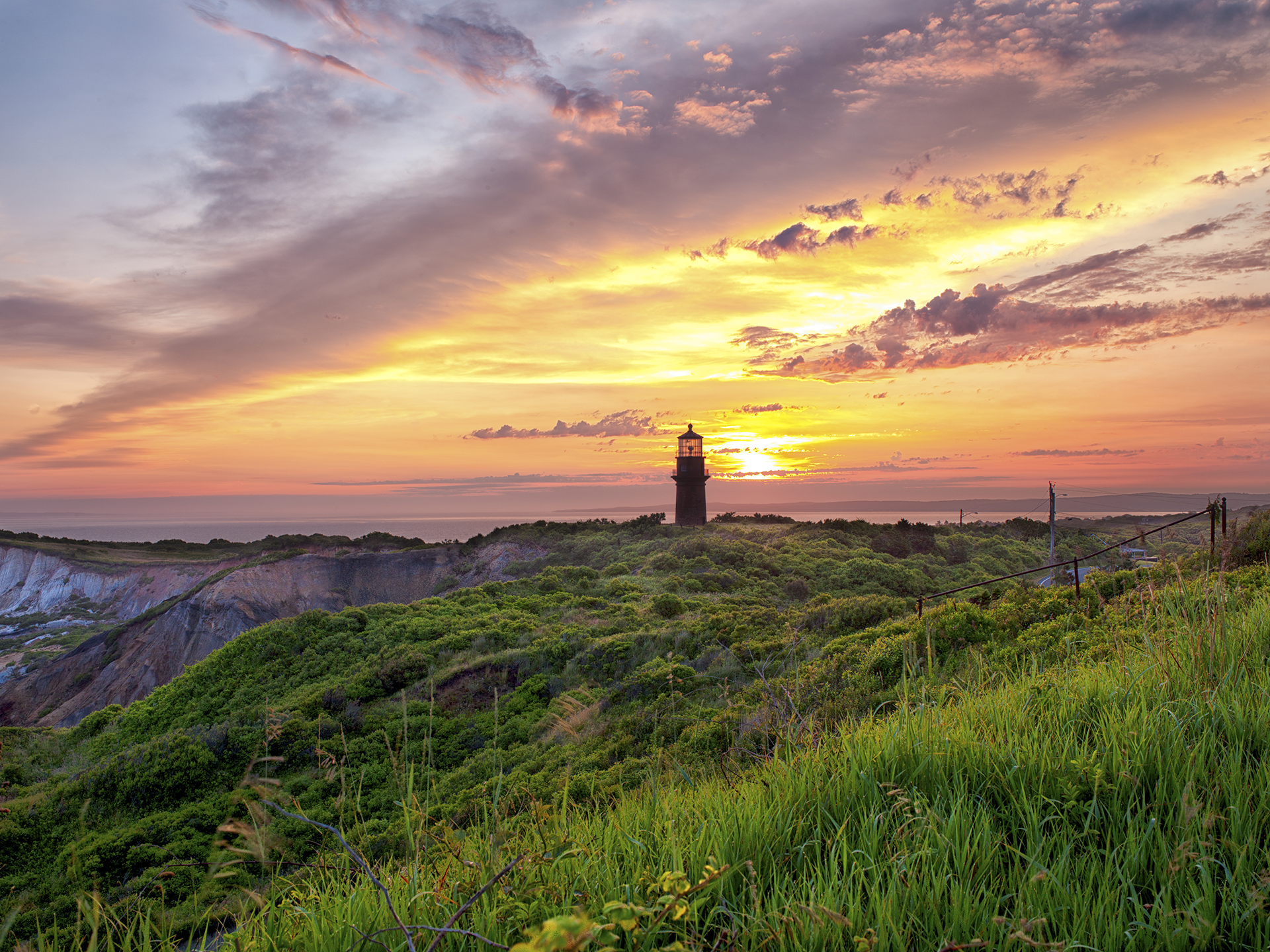 Sunset over a lighthouse on a cliff, with colorful sky and green foliage.