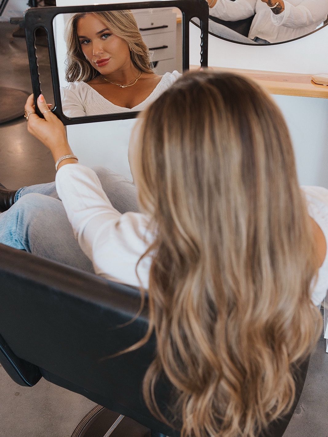 Woman in white top looking at her long wavy blonde hair in a mirror at a salon.