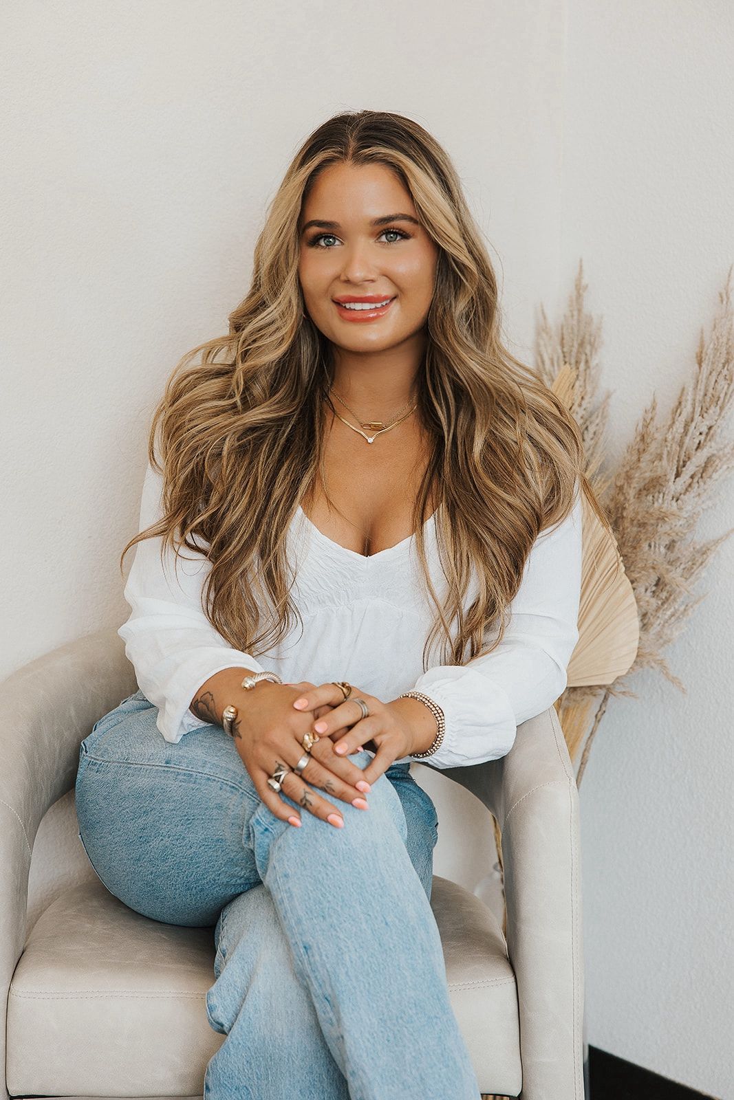 Woman with wavy blonde hair, smiling, seated in a chair, wearing white top and jeans.