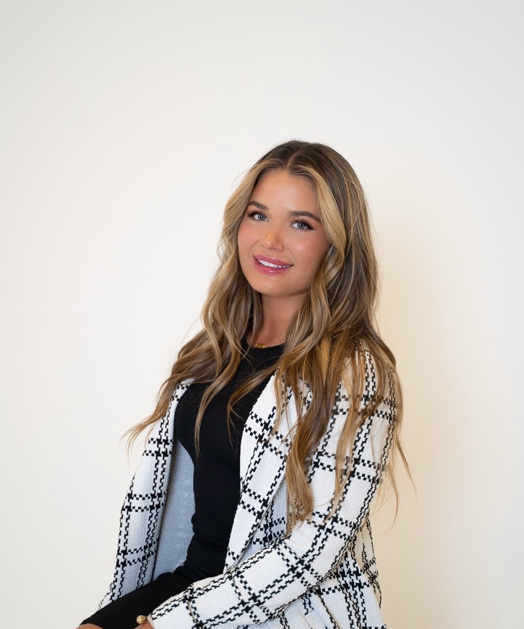 Woman with long wavy hair wearing a black dress and a black and white plaid blazer, smiling against a white background.