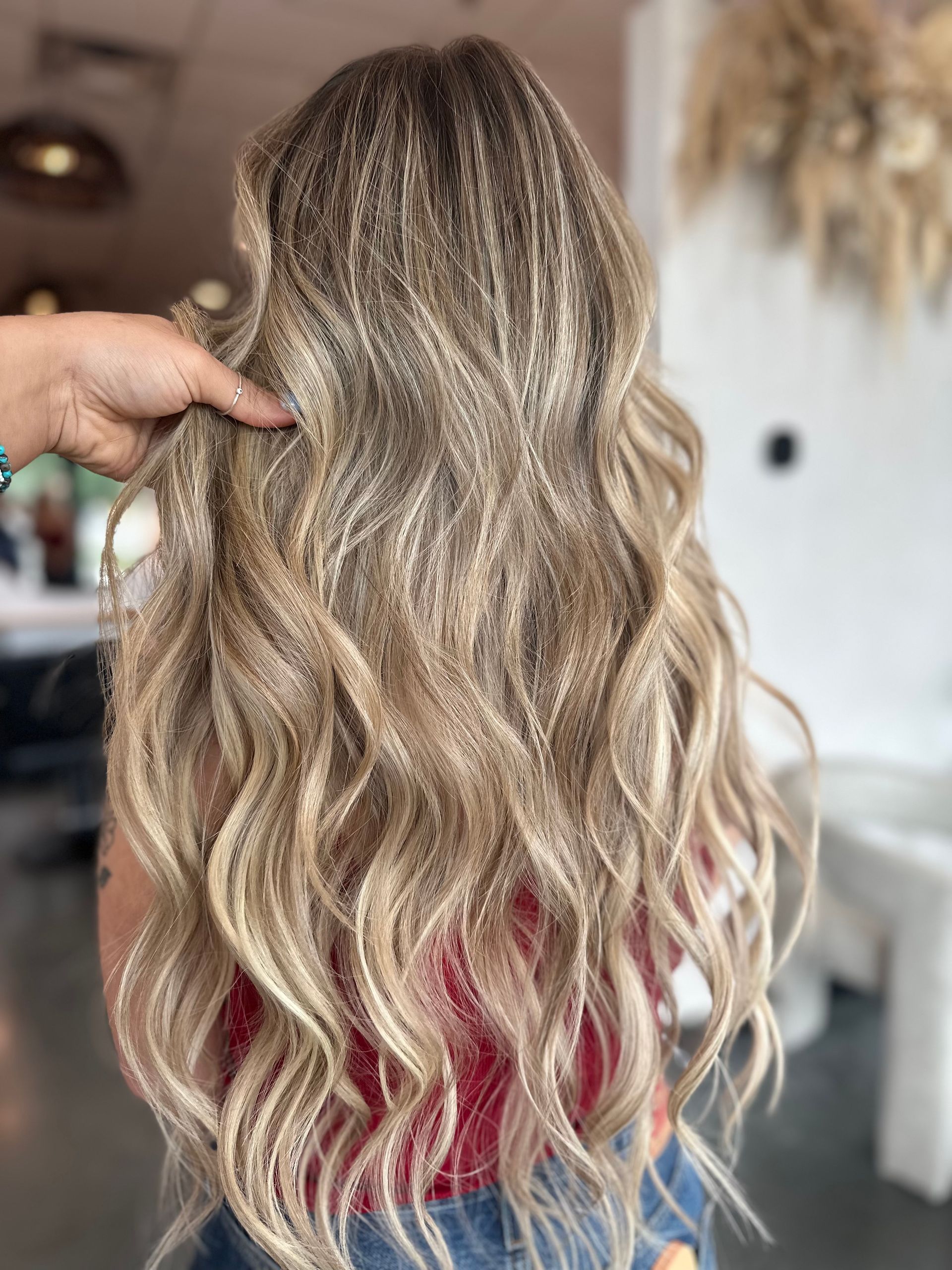 Woman's long, wavy hair; light brown at roots, blonde highlights, hand touching it. Indoors, blurred background.