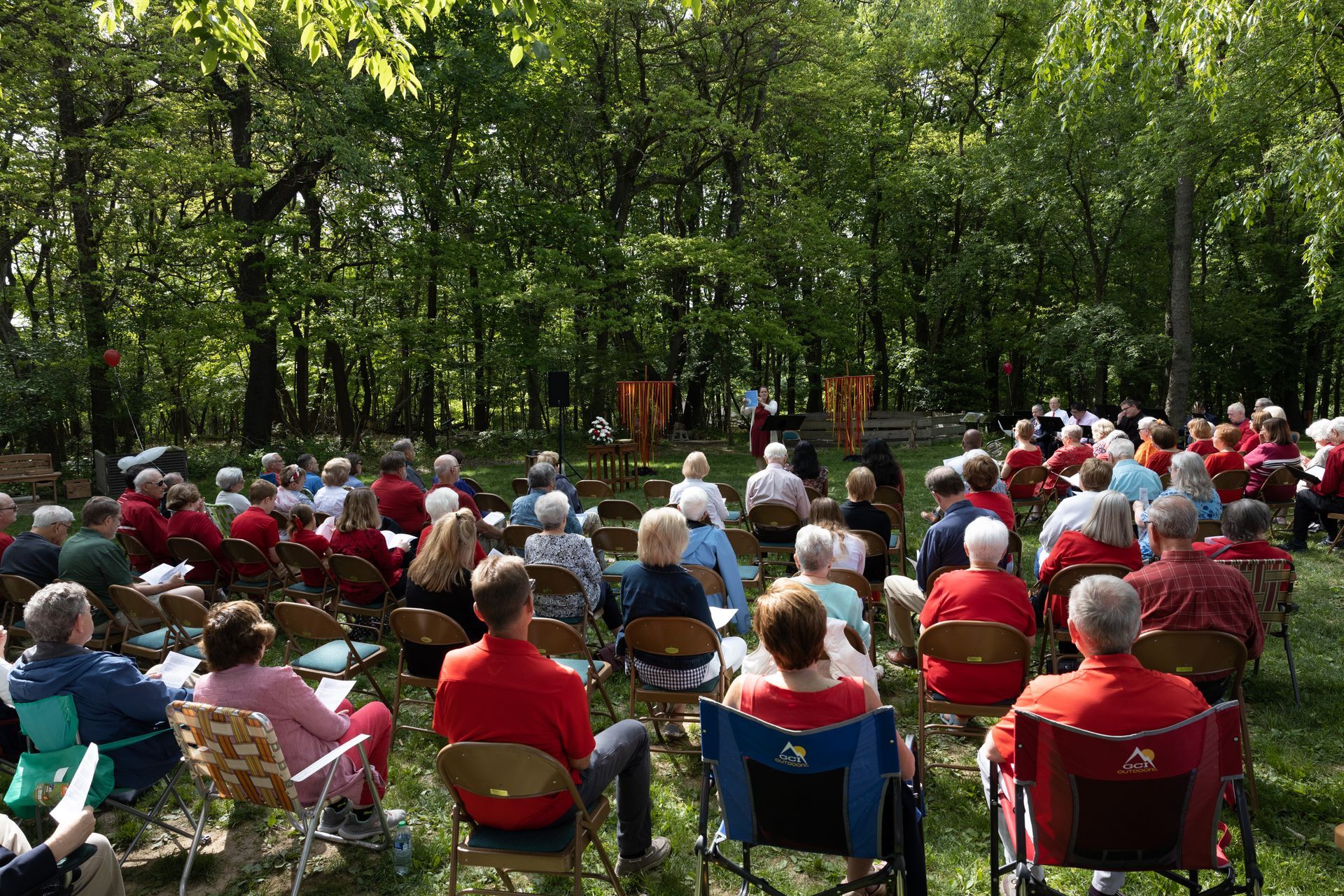 A church congregation gathered outdoors, seated in chairs amidst the woods.