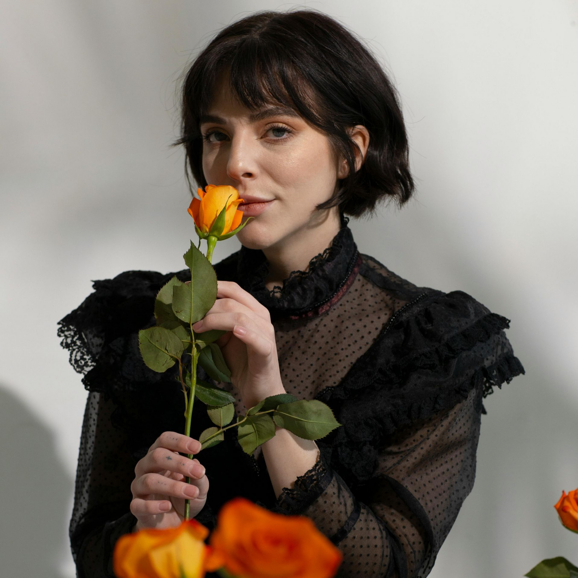 Mujer oliendo una rosa naranja, vestida con un top de encaje negro y jeans, sentada cerca de flores.