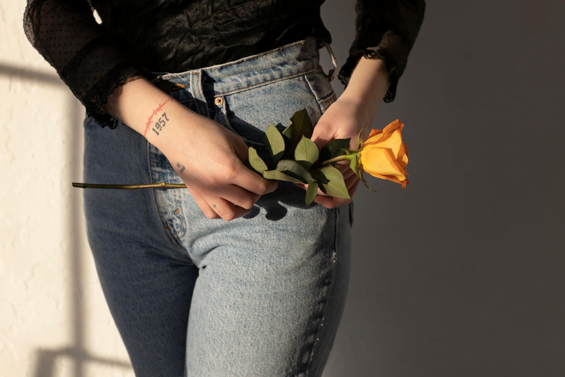 Persona en jeans sosteniendo una rosa amarilla, tatuaje en el brazo.