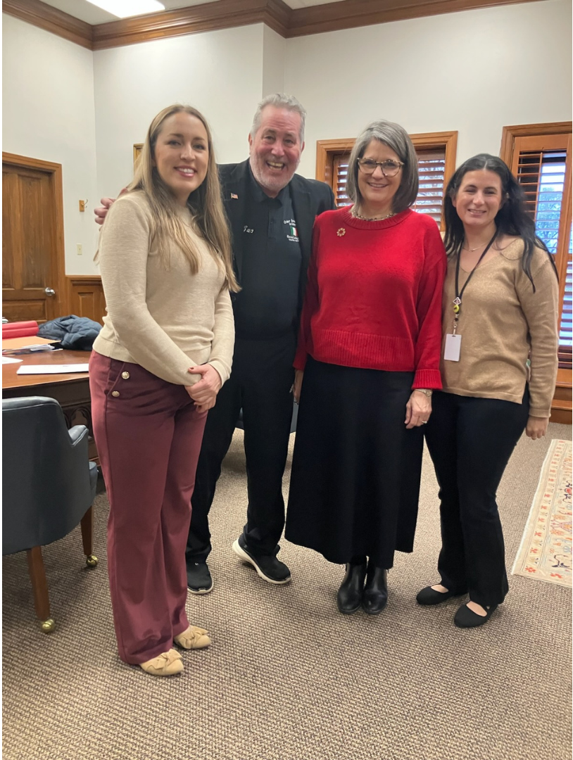 Four people pose in a room. Two women in red and tan tops stand with two men in black. They smile at the camera.