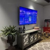 A large TV mounted above a grey mirrored media console, next to a large potted plant and a red popcorn cart.
