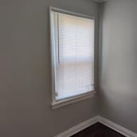 A window with white horizontal blinds in a corner with grey walls and dark hardwood flooring.