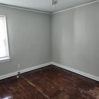 An empty room with gray walls, white trim, a single window on the left, and dark, worn hardwood flooring.