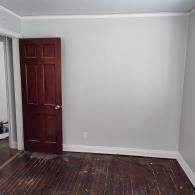 An empty room with light gray walls, a closed dark wood paneled door, and worn hardwood floors.