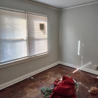 An empty room with light gray walls, white trim, blinds on the windows, and a dark wood floor with some debris.