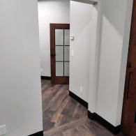 A hallway with gray walls, dark wood-look plank flooring, black baseboards, and a glass-paneled wooden door in the distance.