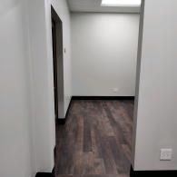 A bright, minimalist office space with dark wood-look flooring, light gray walls, black baseboards, and a doorway.