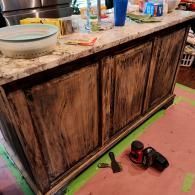 A kitchen island with dark, distressed wood-grain cabinet doors and a light speckled countertop, with tools on the floor.