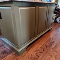 A kitchen island with matte olive green cabinets and a neutral-toned stone countertop, set on a hardwood floor.