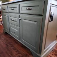A sage green kitchen island with drawers and a cabinet door, featuring silver hardware and a marble countertop.