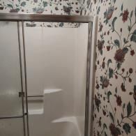 A walk-in shower with a silver-framed sliding door, set against a wall featuring vintage-style floral wallpaper.