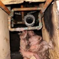 A view looking into a joist bay showing a loop of copper plumbing pipes surrounding a dark pipe drain opening.
