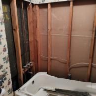 A bathtub in a bathroom during renovation, with wall panels removed to expose the wooden framing and plumbing fixtures.