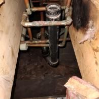 A vertical black plumbing pipe surrounded by a copper water pipe frame between two wooden ceiling joists.