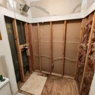 An unfinished bathroom space showing exposed wood wall studs, subflooring, and plumbing pipes behind a shower wall.