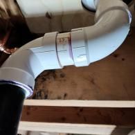 A white PVC plumbing joint connecting two pipes, with visible purple primer, mounted under a wooden subfloor.