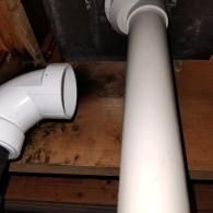 Disconnected white PVC plumbing pipes situated against a wooden surface inside a wall or crawlspace.
