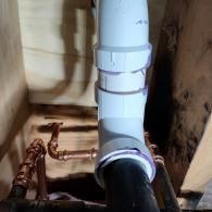 A white PVC drain pipe connected to a black pipe, with copper water supply lines visible in the background under a sink.
