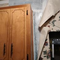 Wooden kitchen cabinets sit next to a wall where floral wallpaper is peeling to reveal a white, textured surface.