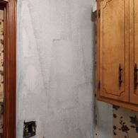 A wall section in a room with a light gray, unevenly painted surface, a wooden cabinet, and remnants of floral wallpaper.