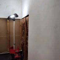 A bright LED work light on a tall red stand illuminates a wall with exposed wooden studs and insulation in a room.