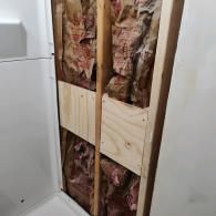 An open wall cavity showing wood framing, pink fiberglass insulation, and newly installed plywood backing for support.