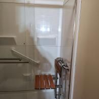 A walk-in shower with white tiled walls, a white grab bar, and a wall-mounted teak wood folding shower seat.