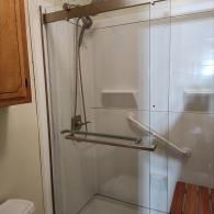 Walk-in shower with a sliding glass door, adjustable handheld shower head, grab bar, and a wooden folding seat.