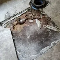 A damaged bathroom floor with cracked tiles, exposed subflooring, and a toilet drain pipe flange in the corner.