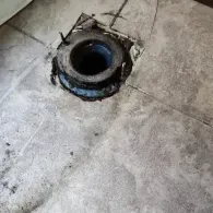 A close-up of a toilet floor flange surrounded by grey tile, with two bolts protruding upwards for toilet installation.