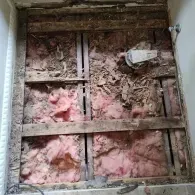 A view of an open floor sub-structure with exposed wooden joists, pink fiberglass insulation, and plumbing components.