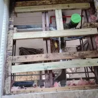 An open ceiling cavity showing wooden floor joists, new support lumber, and exposed insulation and plumbing.