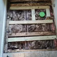 Open wall cavity with exposed wooden framing, fiberglass insulation, and a circular green electrical box.