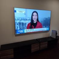 A television mounted on a beige wall above a dark, modern entertainment console, showing a news broadcast with a reporter.