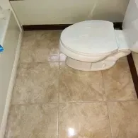 A white toilet positioned in a corner of a bathroom with light brown, marble-patterned floor tiles.