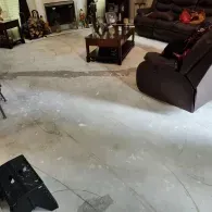 A living room with an exposed concrete floor, brown furniture, a wooden coffee table, and a black footrest in the foreground.