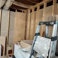 A room under renovation with wooden wall studs, drywall panels, and a silver folding step ladder in the foreground.